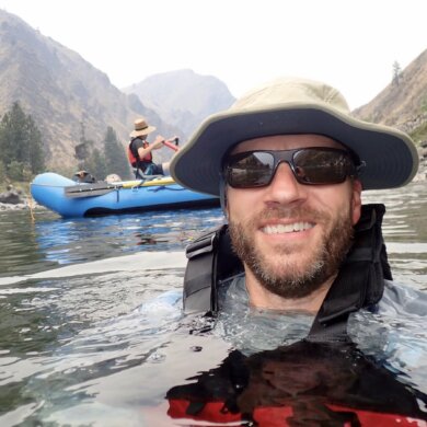 Author wearing Shelta Hat while swimming in river with whitewater raft in background.