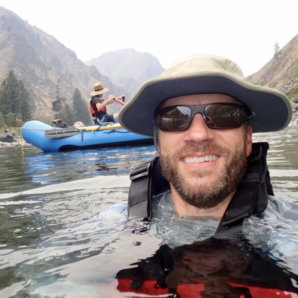 Author wearing Shelta Hat while swimming in river with whitewater raft in background.