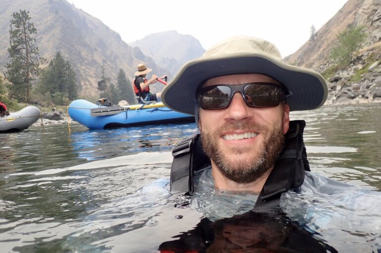 Author wearing Shelta Hat while swimming in river with whitewater raft in background.