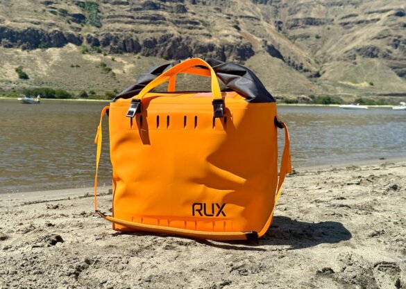 RUX Soft Cooler Bag in the orange color option on a sandy beach in front of water.