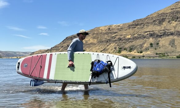 Author carrying a paddleboard while wearing a Firebird V2 sun hat in the sunshine at a beach.
