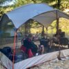 Two people testing a screen shelter while sitting inside of a screen tent in camp chairs next to a camping table.