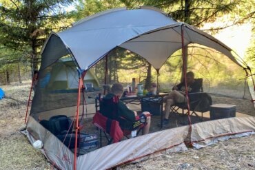 Two people testing a screen shelter while sitting inside of a screen tent in camp chairs next to a camping table.