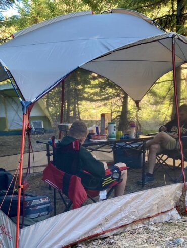 Two people testing a screen shelter while sitting inside of a screen tent in camp chairs next to a camping table.