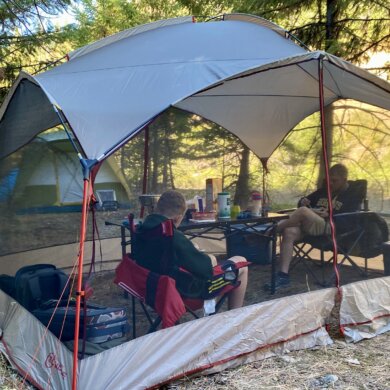 Two people testing a screen shelter while sitting inside of a screen tent in camp chairs next to a camping table.