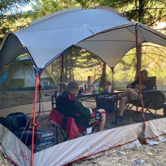 Two people testing a screen shelter while sitting inside of a screen tent in camp chairs next to a camping table.