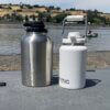 A 1-gallon RTIC Outback Jug in stainless steel next to a white 1/2 gallon RTIC Outback Jug on a paddle board on a beach near a river.