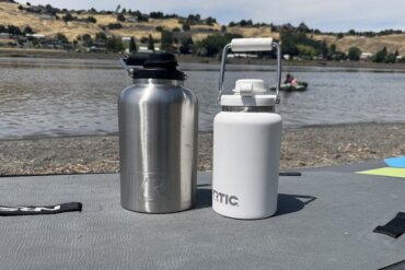 A 1-gallon RTIC Outback Jug in stainless steel next to a white 1/2 gallon RTIC Outback Jug on a paddle board on a beach near a river.