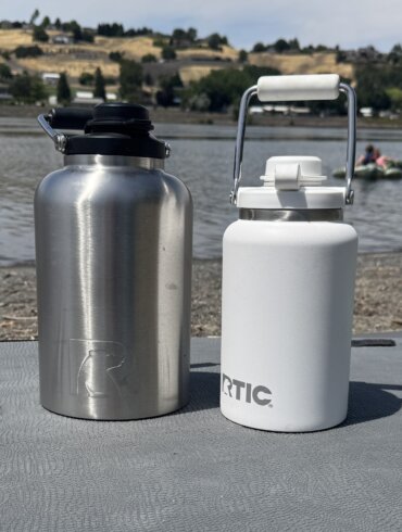 A 1-gallon RTIC Outback Jug in stainless steel next to a white 1/2 gallon RTIC Outback Jug on a paddle board on a beach near a river.