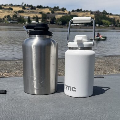 A 1-gallon RTIC Outback Jug in stainless steel next to a white 1/2 gallon RTIC Outback Jug on a paddle board on a beach near a river.