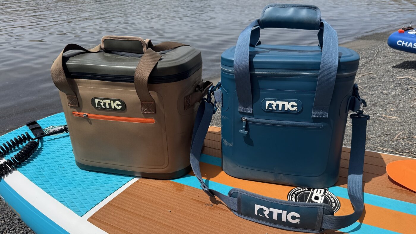 The RTIC 30-can and 20-can Ultra-Tough Soft Cooler side-by-side on an iSUP paddleboard on a beach near water.