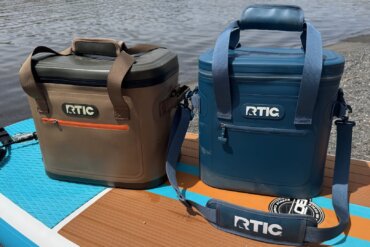 The RTIC 30-can and 20-can Ultra-Tough Soft Cooler side-by-side on an iSUP paddleboard on a beach near water.