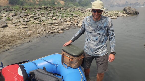 The author standing in a river next to the RTIC Ultra-Tough Soft Cooler 30 can option, near a whitewater raft.