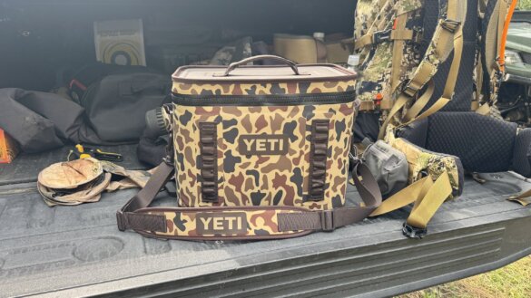 The author's YETI Hopper Flip 18 Soft Cooler in the Woodlands Camo color pattern sits on a tailgate next to a hunting backpack and outdoor gear.